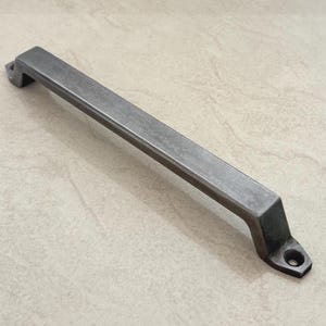 Pode incluir: Um puxador de armário retangular de metal cinza com acabamento texturizado. A alça tem um design plano em forma de barra com suportes angulados e um único orifício de montagem em cada extremidade. Ferragens para móveis.