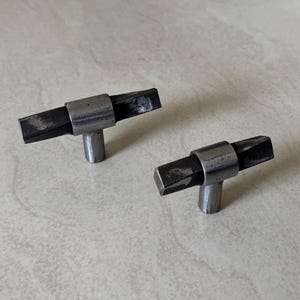 May include: Two metal cabinet knobs with a T-bar design. The knobs have a cylindrical base and a rectangular bar with beveled edges. The metal has a dark, textured finish, suggesting a rustic or industrial style. The knobs are likely for drawers or cabinets.