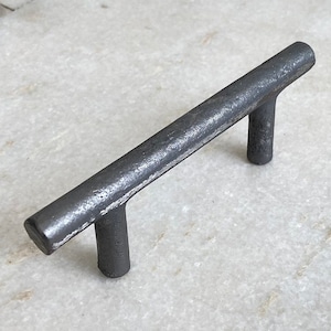May include: A black metal cabinet pull with a simple, rectangular design. The pull has a slightly distressed finish, giving it a rustic look.