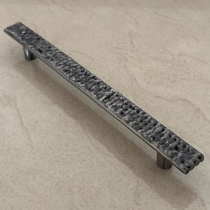 Könnte beinhalten: Ein rechteckiger, strukturierter, silberfarbener Schrankgriff mit zwei zylindrischen Pfosten. Der Griff hat ein gehämmertes Aussehen und verleiht ihm eine rustikale Note. Die Hardware ist für Möbel und Schränke konzipiert.