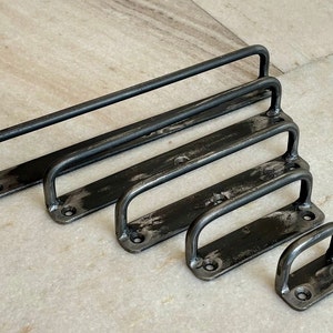 Puede incluir: Cinco tiradores de cajón de metal negro de diferentes tamaños. Los tiradores son todos de forma rectangular con bordes redondeados y una sola manija en el centro.