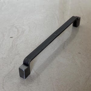 May include: A dark gray metal cabinet pull with a rectangular handle and square ends. The handle is straight and slightly curved at the ends. The hardware is designed for use on drawers or cabinet doors.