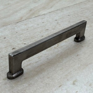 Op de afbeelding: Een zilveren metalen kastgreep met een rechthoekige vorm en afgeronde randen.