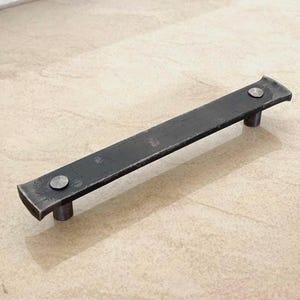 Könnte beinhalten: Ein dunkelgrauer, rechteckiger Metallschrankgriff mit abgerundeten Enden und zwei zylindrischen Pfosten. Der Griff ist mit zwei silbernen Nieten befestigt. Die Hardware ist für Schränke und Schubladen konzipiert.