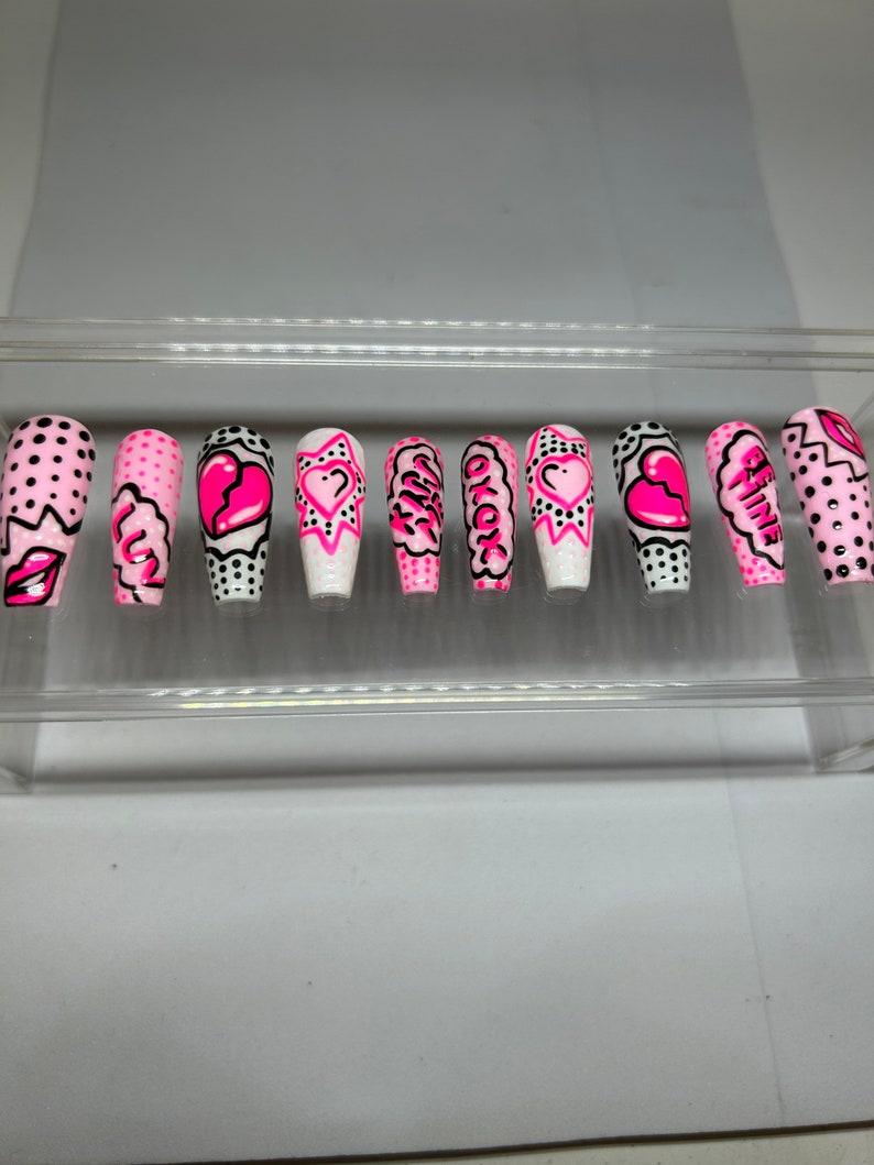 May include: Set of ten pink and white false nails with a comic book theme. The nails feature hearts, stars, and words like "Girl" and "Be Mine".