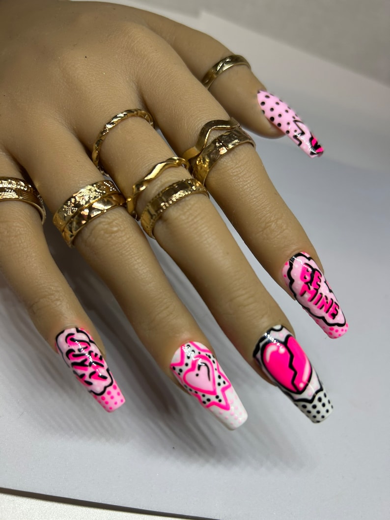 May include: Pink and white long press-on nails with black and pink designs. The designs include hearts, stars, and the words "Be Mine" and "Slay".