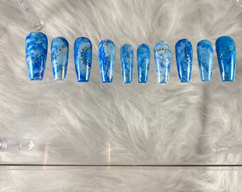 May include: A set of ten blue and white marble-effect press-on nails. Each nail is a different size and features gold glitter accents. The nails are arranged on a clear, rectangular display.