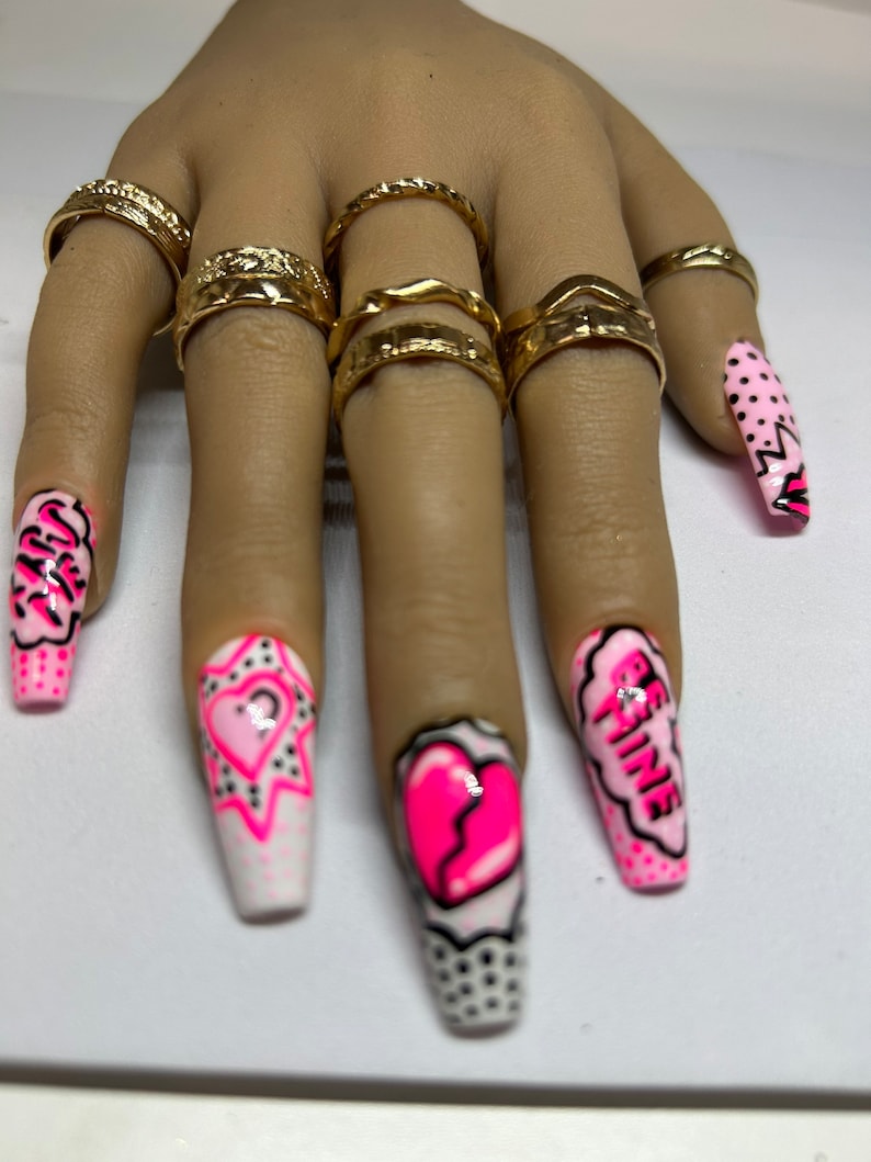 May include: Pink and white long false nails with different designs. One nail has a pink heart with a crack in it, another has a pink heart with a question mark, another has the words "Be Fine" in a pink speech bubble, and another has a pink and white polka dot design.