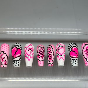 May include: Set of ten pink and black false nails with various designs. The designs include hearts, polka dots, and words like "Girl" and "Be Mine".
