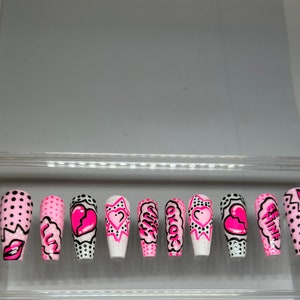 May include: Set of ten pink and white false nails with different designs. Some nails have black polka dots, a broken heart, a star, and the words "Okay" and "Be Fine".