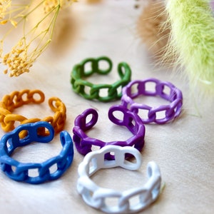 May include: Six colourful chain link rings in yellow, green, purple, blue, and white. The rings are arranged on a light beige surface.