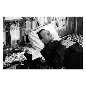 K&ouml;nnte beinhalten: Schwarzwei&szlig;foto eines Mannes, der im Bett liegt. Er tr&auml;gt einen Anzug und hat die H&auml;nde auf der Brust gefaltet. Auf einem Tisch neben dem Bett stehen Gl&auml;ser und Geld.