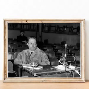 Könnte beinhalten: Schwarzweißfoto eines Mannes, der auf einem Stuhl sitzt und ein Glas Flüssigkeit vor sich hat. Er trägt eine Sakkojacke und eine Krawatte. Vor ihm befindet sich ein Mikrofon und auf dem Tisch eine Flasche Flüssigkeit.