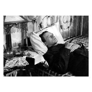 K&ouml;nnte beinhalten: Ein Schwarzwei&szlig;foto eines Mannes, der mit besorgtem Gesichtsausdruck im Bett liegt. Er tr&auml;gt einen Anzug und h&auml;lt eine Zigarette in der Hand. Auf einem Tisch neben dem Bett stehen Gl&auml;ser mit Alkohol und Geld.