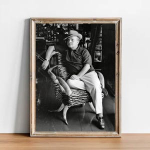May include: Black and white framed photograph of a person seated in a chair. The chair has a zebra print seat and curved armrests. The person is wearing a hat, polo shirt, and pants. The photograph is displayed in a wooden frame.
