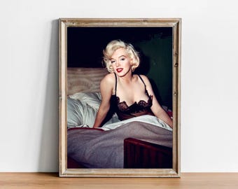 May include: A framed photographic print featuring a blonde woman with red lipstick wearing a black lace camisole, positioned in a bed with white pillows and a grey blanket. The image is encased in a light-colored wooden frame with a visible grain.