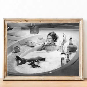 May include: Black and white photograph of a woman in a bathtub full of bubbles. She is on the phone and has a variety of bottles and toiletries around her.
