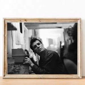 Puede incluir: Fotografía en blanco y negro enmarcada de un hombre mirando hacia arriba, apoyado en una máquina. La fotografía está en un marco de madera de color dorado, con un fondo borroso.