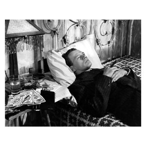 K&ouml;nnte beinhalten: Schwarzwei&szlig;foto eines Mannes, der im Bett liegt. Er tr&auml;gt einen Anzug und hat die H&auml;nde auf der Brust gefaltet. Auf einem Tisch neben dem Bett stehen Gl&auml;ser und Geld.