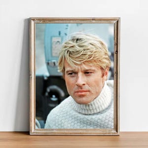 May include: Framed portrait of a man with fair hair wearing a white turtleneck sweater. The portrait is in a wooden frame and is set against a light blue background. The man is looking towards the viewer.