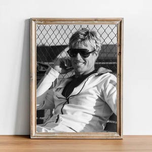May include: Black and white framed photograph of a smiling person wearing sunglasses and a white jumpsuit. The person is sitting in front of a chain-link fence. The photograph is in a wooden frame.