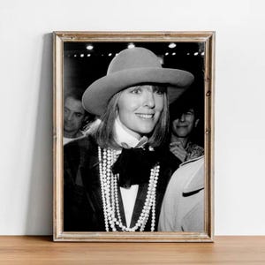 Puede incluir: Fotografía enmarcada en blanco y negro de una mujer con sombrero de ala ancha, blazer oscuro y collar de perlas. La mujer sonríe, y hay otras personas en el fondo.