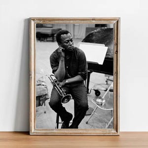 May include: Black and white photo of a man sitting on a stool, holding a trumpet. He is wearing a dark shirt and jeans. The photo is framed in a wooden frame.
