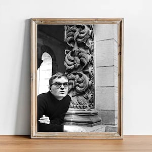 May include: Black and white framed photograph featuring a man wearing glasses and a black turtleneck, leaning against a decorative stone pillar. The photograph is set in an architectural space with ornate details.