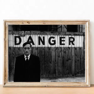 May include: Black and white photograph of a man standing in front of a wooden fence with the word "DANGER" painted in black letters on a white background.