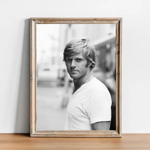 May include: Black and white framed portrait of a man with light-colored hair and a white t-shirt. The portrait is in a wooden frame and is displayed on a wooden surface.