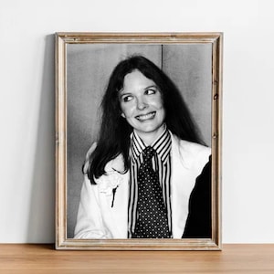 Puede incluir: Retrato en blanco y negro enmarcado de una mujer sonriendo. Lleva una camisa a rayas, una corbata de lunares y una chaqueta blanca. El retrato está en un marco de madera, expuesto sobre una superficie de madera.