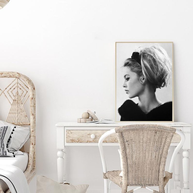 May include: Black and white photograph of a woman with her hair styled in a bouffant, wearing a fur coat. The photo is framed and hanging on a white wall above a white desk with a wicker chair.