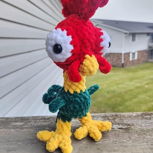 May include: A handmade crochet rooster toy with a vibrant color scheme. The rooster features a red head and crest, white and black eyes, a yellow beak and feet, and a green body. The plush toy is standing on a wooden surface.