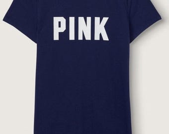 Può includere: Una t-shirt a maniche corte blu navy con la parola PINK stampata in grandi lettere maiuscole bianche sul petto. Il capo presenta uno scollo rotondo e una piccola etichetta bianca del marchio stampata all'interno del colletto.