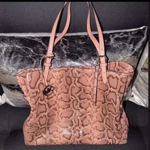 May include: A pink and brown snakeskin-patterned tote bag with matching handles and a gold-tone logo charm. The bag features a spacious interior and is designed for everyday use.