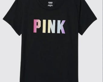 Può includere: Una t-shirt nera a maniche corte con scollo rotondo e la parola PINK stampata sul petto in una sfumatura di colori che va dal rosa al giallo, azzurro e viola. L'etichetta interna sul collo riporta anch'essa la parola PINK.