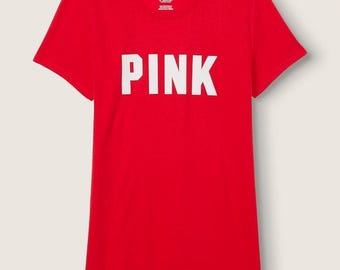 Può includere: Una t-shirt rossa a maniche corte con la parola PINK stampata in grandi lettere maiuscole bianche sul petto. L'interno del colletto presenta una piccola etichetta bianca con il testo PINK stampato in nero, insieme ad altro testo.
