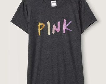 Può includere: Una t-shirt a maniche corte color grigio antracite con scollo rotondo. La parte anteriore presenta la scritta PINK stampata in un carattere multicolore, glitterato e in stile pennellata con tonalità gialle, arancioni, rosa e viola. Un'etichetta bianca è sul collo.