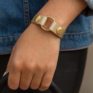 May include: A gold-coloured leather bracelet with a circular gold ring and gold snap closures. The bracelet is worn on a wrist, and the person is holding a black handbag.