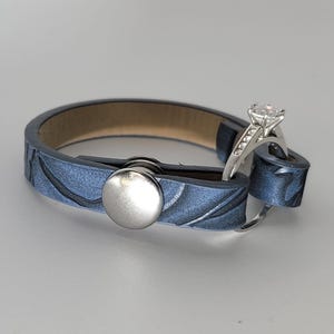 May include: A blue leather bracelet featuring a textured surface with a silver-toned metal snap button closure. A silver ring with a clear stone setting is looped through the end of the bracelet, secured by the snap button mechanism.