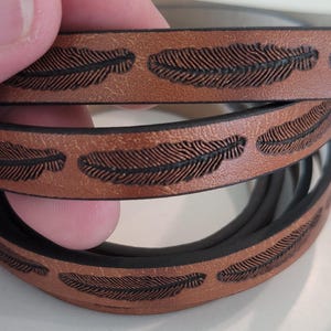 May include: Close-up of a brown leather bracelet with a repeating feather design. The feathers are dark brown and etched into the leather. The bracelet has a black interior.