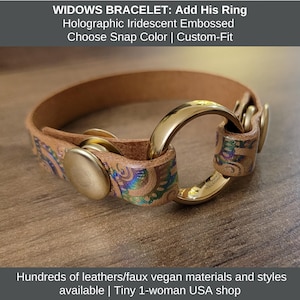 May include: A tan leather bracelet with a gold-colored ring and snap closure. The bracelet features holographic iridescent embossed detailing. The text "WIDOWS BRACELET: Add His Ring" is visible at the top of the image.