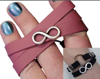 Op de afbeelding: Een wikkelarmband met een zilveren oneindigheidssymbool en een druksluiting. De armband is stofroze van kleur en wikkelt zich om de pols. De tekst op de afbeelding luidt "RING HOLDER BRACELET" en "Choose Snap Color".