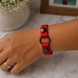 May include: A bright red nylon wristband featuring two matte black snap buttons and a central black metal ring. The strap is constructed from durable woven fabric, secured with the snap closures, and centered with a circular metal hardware component.