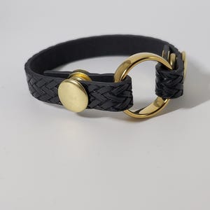 Peut inclure: Un bracelet en cuir tressé noir doté d'un anneau en métal doré poli et d'une fermeture à bouton-pression assortie. La bande présente un motif tissé texturé et est fixée par une attache métallique circulaire sur un côté.