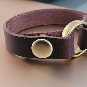 May include: A brown leather bracelet with a gold metal ring and a gold metal button closure.