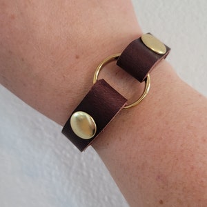 May include: A brown leather bracelet with a gold metal ring and two gold metal snaps.
