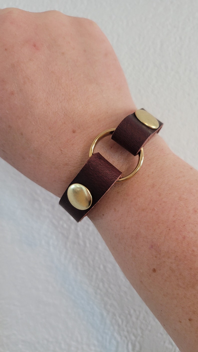 May include: A brown leather bracelet with a gold metal ring and two gold metal snaps.
