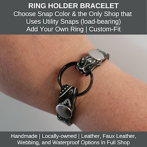 May include: A black and white patterned ring holder bracelet with a large black ring. The bracelet has silver snaps and is designed to hold a ring. The text on the image says "RING HOLDER BRACELET".