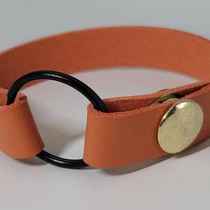 May include: Orange leather bracelet with a black ring accent and gold-coloured button closures. The bracelet is a simple, modern design, suitable for everyday wear. The bracelet is approximately 2 cm wide.